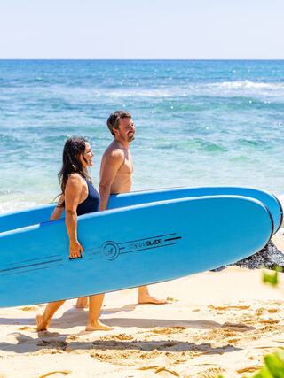 A couple walks along a sunny beach, carrying blue surfboards, with calm waves and a clear sky in the background.