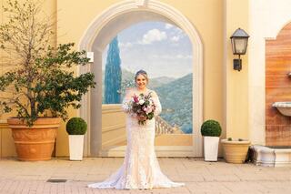 A bride in a stunning lace gown holds a bouquet, smiling against a picturesque mural of mountains, surrounded by greenery and elegant decor.