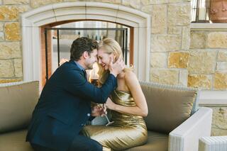 A couple shares an intimate moment, playfully leaning in for a kiss, surrounded by a cozy setting with a stone fireplace.