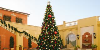 A festive Christmas tree adorned with colorful ornaments stands in a beautifully decorated courtyard with lush greenery and festive garlands.