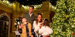 A joyful family stands together in the snow, surrounded by holiday lights and a decorated tree, sharing laughter and warm moments.