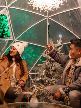 A festive couple enjoys drinks inside a cozy igloo-like space, surrounded by a decorated Christmas tree and colorful lights.