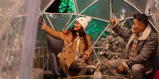 A couple enjoys a cozy moment inside a transparent dome, taking selfies near a decorated Christmas tree and festive lights.