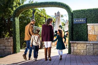 A family of four walks through a lush archway, enjoying a sunny day, with the entrance to a vibrant venue in the background.