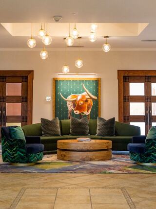 A stylish lounge features a green sectional sofa, plush chairs, warm lighting, and a vibrant bull painting as a focal point.