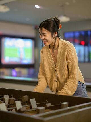 A young woman in a cozy cardigan enjoys a game while smiling, with vibrant background lights and a TV showing sports behind her.