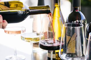 A hand pours rich red wine into a glass, surrounded by various wine bottles and glasses, creating a warm and inviting atmosphere.