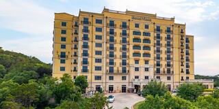 A tall, yellow hotel building surrounded by lush greenery, featuring multiple balconies and a welcoming entrance.