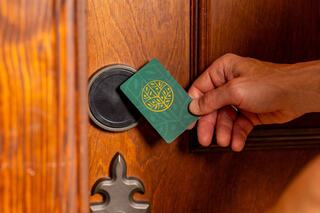 A hand holds a key card near a door lock, preparing to unlock it, with a wooden door in the background.