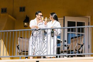 A couple in white robes clinks champagne glasses on a balcony, enjoying a joyful moment together in the sun.