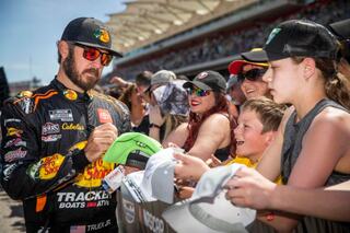 A race car driver signs autographs for enthusiastic fans, including children, at a lively event under a clear blue sky.