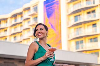 A woman in a green dress smiles while holding a cocktail, with a colorful hotel facade in the background.