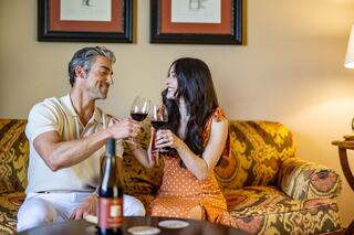 A couple clinks wine glasses while smiling at each other, surrounded by a cozy, stylish living space with warm lighting.