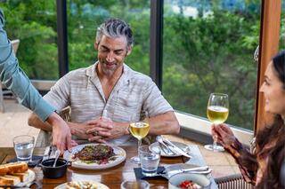 A man enjoys a meal being served at a table, while a woman seated nearby raises her glass, all set against a lush outdoor backdrop.