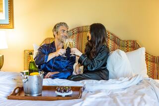 A couple in elegant pajamas enjoy a champagne toast in bed, surrounded by a cozy atmosphere and a tray of treats.