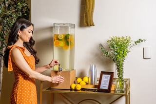 A woman in a polka dot dress dispenses refreshing citrus-infused water from a stylish beverage dispenser on a gold cart adorned with lemons and flowers.