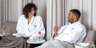 A man and woman in spa robes share a light moment, enjoying drinks and a cozy atmosphere, surrounded by elegant decor and soft lighting.