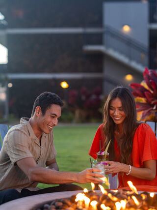 A joyful couple shares drinks by a glowing fire pit, surrounded by greenery and warm evening lights, radiating a cozy, intimate atmosphere.