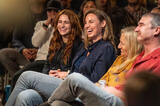 Two women share a joyous moment, laughing in a lively crowd. Others around them also enjoy the humor of the event.