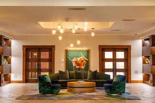 A stylish lobby with a green sofa, patterned chairs, warm lighting, and wooden shelves, featuring a vibrant art piece of a bull.