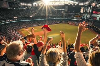 A lively crowd at a baseball stadium cheers enthusiastically, raising their arms and flags under bright sunlight. Excitement fills the air.