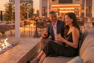 A couple enjoys drinks by a cozy fire, sharing smiles in a chic rooftop setting at sunset, surrounded by elegant decor and ambiance.