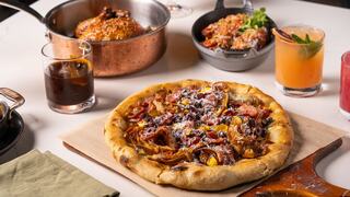 A delicious spread featuring a hearty pizza topped with meats and corn, accompanied by cocktails and side dishes in an inviting setting.
