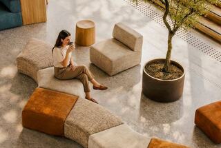 A woman enjoys a drink while seated on modern, textured furniture amidst a bright, open space featuring greenery.