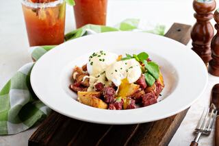 A plate features a hearty dish with poached eggs, corned beef, and potatoes, garnished with chives and greens, alongside refreshing drinks.