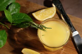 A glass of yellow juice garnished with a sprig of rosemary, accompanied by lemon wedges and green leaves on a wooden surface.
