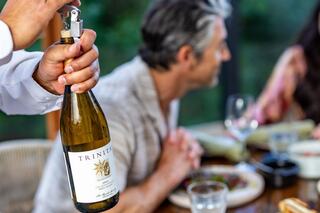 A server pours wine from a bottle labeled "Trinité" while guests share a meal, enjoying a warm, inviting atmosphere.