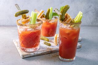 Three vibrant Bloody Mary cocktails adorned with celery sticks, pickles, and a spicy rim, presented on a textured surface.