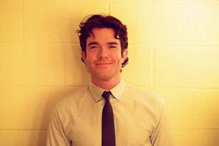 A smiling man with curly hair stands against a light-colored wall, dressed in a light shirt and tie, exuding friendliness and warmth.