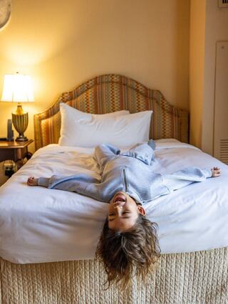 A joyful child lies on a hotel bed, arms spread wide, with a big smile, exuding carefree happiness in a cozy room.
