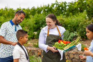 A smiling chef holds a basket of fresh vegetables while engaging with a man and two children in a lush garden setting.