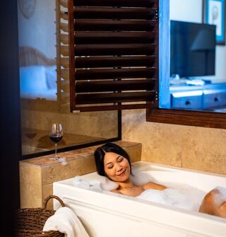 A woman relaxes in a bubble-filled bathtub, enjoying a serene moment with a glass of wine nearby and a cozy atmosphere.