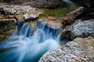 A serene stream flows over smooth rocks, creating a gentle cascade of water amidst a natural landscape of earthy tones and greenery.