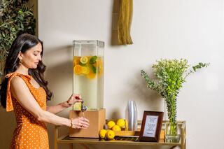A woman in a polka dot dress serves infused water with lemons and herbs from a stylish dispenser, surrounded by fresh flowers and lemons.