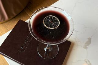 A dark cocktail garnished with a dried lemon slice sits atop a brown menu, contrasting with a light marble surface.