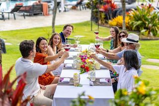 A joyful group toasts over a beautifully set table adorned with flowers, enjoying drinks amidst a vibrant, lush garden setting.