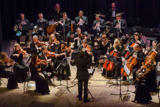 A full orchestra performs on stage, featuring various instruments and musicians in formal attire, focused on the conductor.
