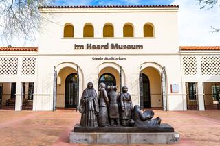 A stone sculpture of figures stands in front of the Heard Museum, showcasing indigenous culture, with a bright, welcoming facade.